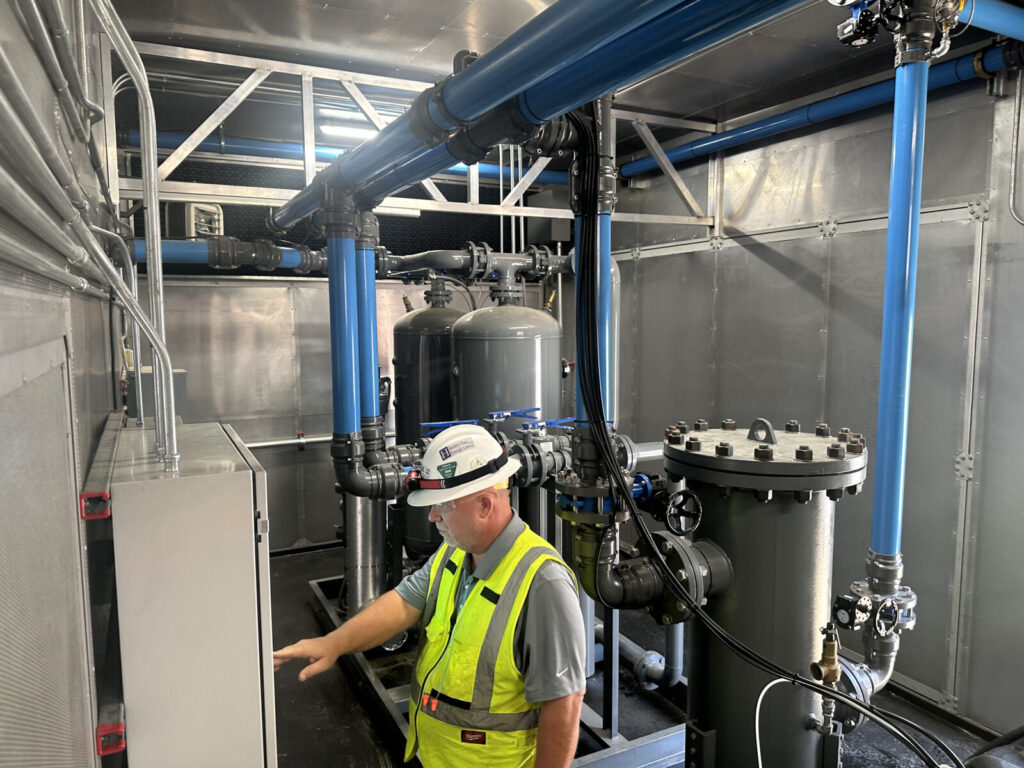 Worker in safety vest and helmet examines control panel in an industrial room with blue pipes and metal tanks.