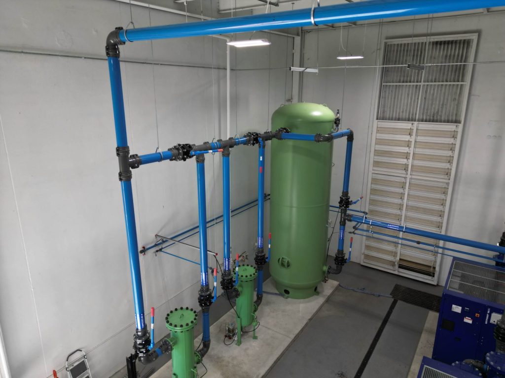Green industrial tank connected to blue pipes inside a clean facility with white walls and overhead lighting.