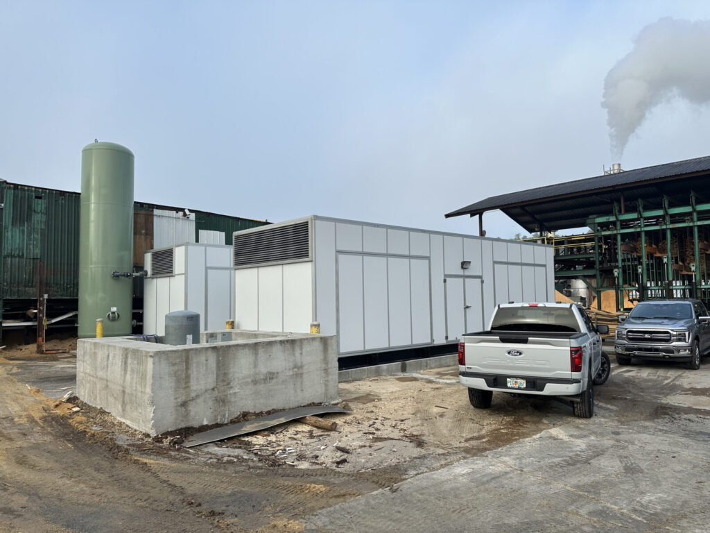 A large white industrial unit sits next to a green tank, parked trucks, and factory buildings with smoke in the background.