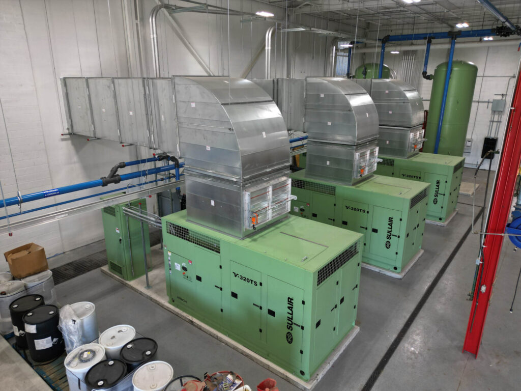 Three large green industrial machines with metal ductwork in a clean, organized facility with barrels and piping nearby.