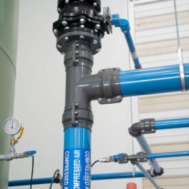Blue compressed air pipes with labeled arrows, connected by gray fittings and valves, in an industrial setting.