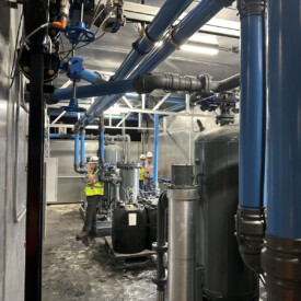 Two workers in safety vests inspect equipment and blue pipes inside an industrial facility with tanks and machinery.