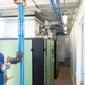 Industrial equipment with blue overhead pipes and green machinery inside a brightly lit, narrow utility room.