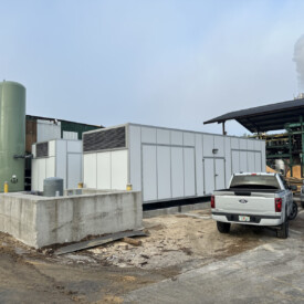 A large white industrial unit sits next to a green tank, parked trucks, and factory buildings with smoke in the background.