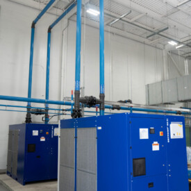 Two large blue industrial machines with vertical blue pipes inside a white-walled facility with a high ceiling.