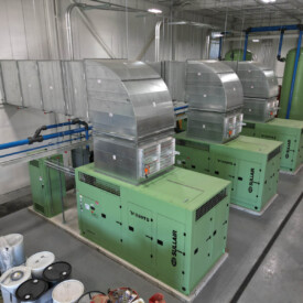 Three large green industrial machines with metal ductwork in a clean, organized facility with barrels and piping nearby.